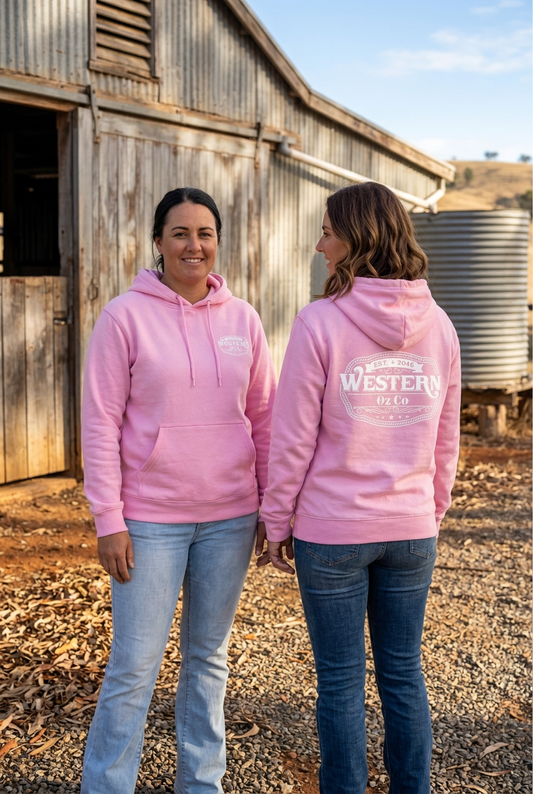 Two women showing the front and back of baby pink Western Oz Co Legends hoodies with white logos outside a rustic shed